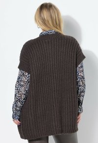 Donna bionda che indossa un cardigan senza maniche a maglia spessa di colore marrone scuro sopra una camicia a maniche lunghe con motivo, vista da dietro.