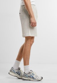 Pantalones cortos de rayas de color claro con una cinturilla elástica, cordón y bolsillo lateral, confeccionados en una tela texturizada. Combinados con zapatos deportivos plateados.