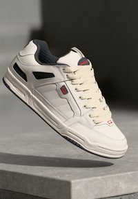 Cream-colored sneaker with textured leather, grey accents, and black mesh panels. Features a cushioned sole and contrasting blue rubber outsole.