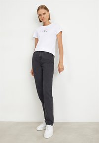 Biela bavlnená tričko s strieborným logom "ck", zkombinované s tmavosivými rifľami a bielymi teniskami, stojace na neutrálnom pozadí.