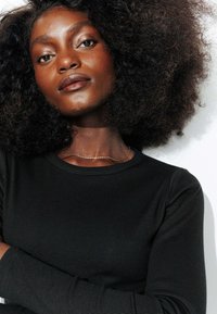 Chemise noire à manches longues côtelées avec un col rond. Un fin collier en chaîne dorée met en valeur le décolleté, complétant le teint de peau foncé.