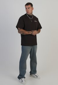 Sort bomulds t-shirt med rund hals og korte ærmer, parret med slidte blå jeans og hvide sneakers. Bemærkelsesværdige tatoveringer synlige på armene.