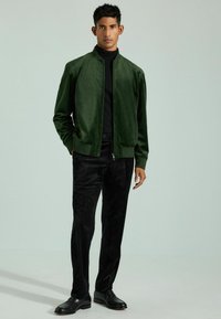 Giacca bomber verde con una superficie liscia, chiusura con zip e polsini a costine, abbinata a pantaloni neri con texture e scarpe nere.