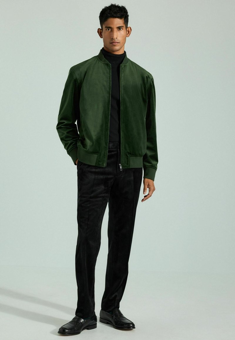 Giacca bomber verde con una superficie liscia, chiusura con zip e polsini a costine, abbinata a pantaloni neri con texture e scarpe nere.