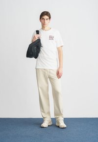 White cotton t-shirt with printed text, beige sweatpants, and cream sneakers. Black shoulder bag with zipper resting on left shoulder.