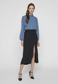 Niebieska bluzka w wzór chevron z falbaniastym dekoltem, zestawiona z czarną spódnicą z wysokim rozcięciem i czarnymi szpilkami nosa.