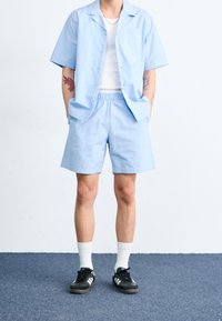 Světle modrá košile s krátkým rukávem a odpovídající šortky, obojí vyrobené z texturované látky. Košile má kapsu a šortky elastický pas.