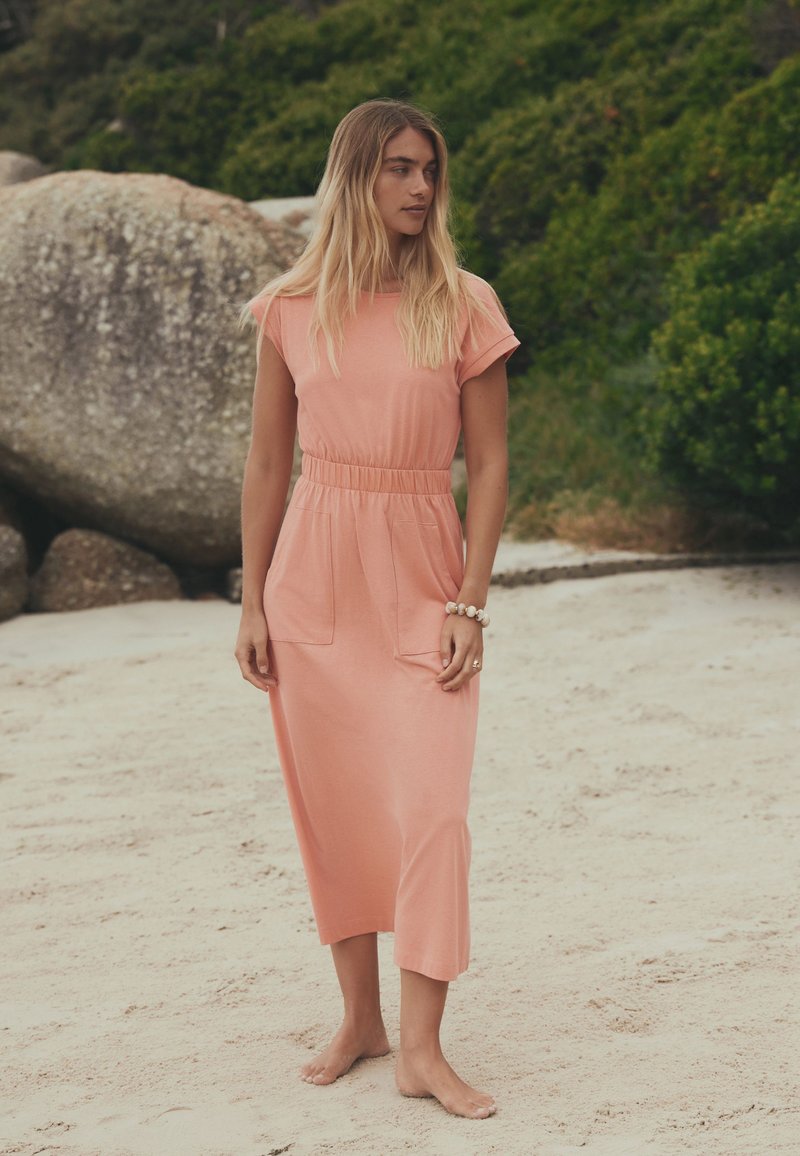 Femme aux longs cheveux blonds, debout pieds nus sur une plage de sable, portant une robe midi rose corail avec des poches et un bracelet.