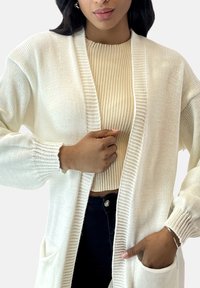 Cardigan bianco lavorato a maglia con apertura frontale, maniche lunghe e tasche. Indossato sopra un top beige aderente a coste, che evidenzia un look stratificato.