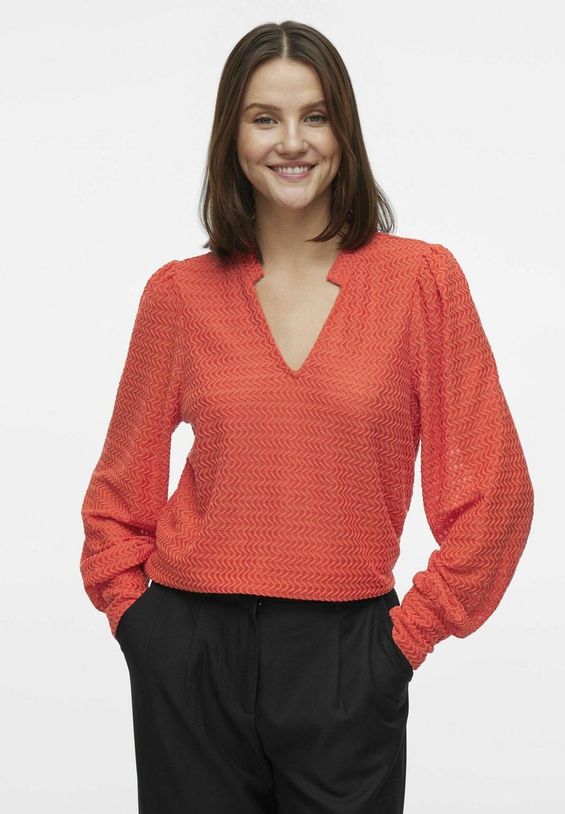 Rød tekstureret bluse med V-hals, lange ærmer og samlede manchetter, båret med sorte bukser. Zigzag mønster synligt på stoffet.