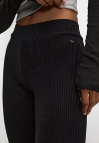 Leggings noirs à côtes avec une large ceinture et un petit détail logo en métal. Le tissu semble extensible et texturé, épousant étroitement le corps.