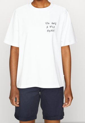 Person trägt ein weißes T-Shirt mit dem Text "You have a nice heart" auf der Brust und dunkelblaue Shorts.