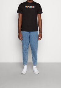 Svart bomulls t-shirt med flerfärgad text "Primitive", matchad med ljusblå denimjeans och vita hög-top sneakers.