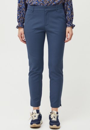 Mujer vistiendo pantalones azul ajustados, blusa azul con estampado y zapatillas azul marino con cordones blancos, de pie frente a un fondo liso.