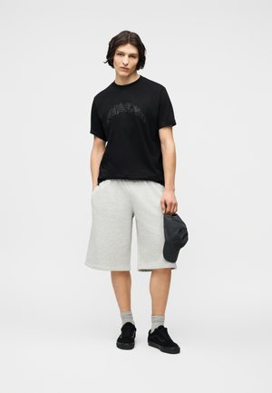 Tânăr purtând un tricou negru cu imprimeu grafic, pantaloni scurți gri deschis până la genunchi, șosete gri, adidași negri, ținând o șapcă închisă la culoare în mâna dreaptă.