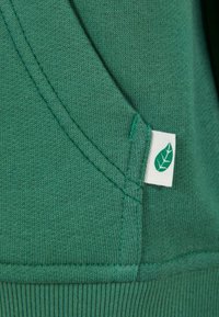 Tessuto di felpa verde in cotone con orlo a coste e una piccola etichetta bianca con un design di foglia verde. Texture soffice e dettagli di cucitura puliti.