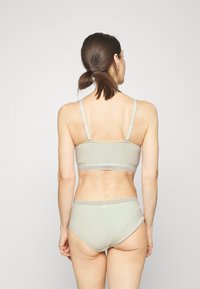 Bralette verde claro com texturas em nervuras, com alças finas e detalhes em renda, emparelhada com cuecas de cintura alta combinantes, vista por trás.