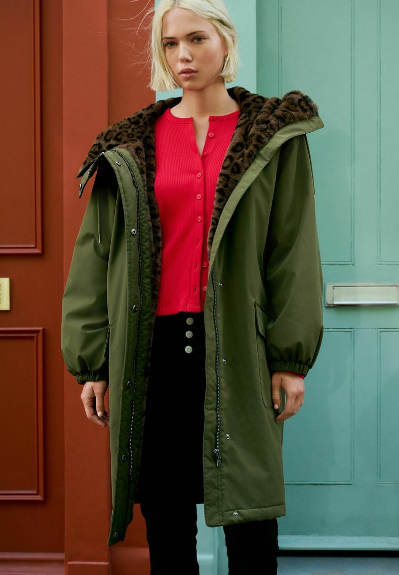 Femme aux cheveux blonds courts portant un parka vert ouvert avec une doublure en imprimé léopard, un pull rouge à boutons et un pantalon noir, se tenant près de portes colorées.