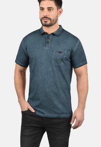 Mørkeblå polo skjorte med krave, tre brune knapper, en brystlomme og en falmet tekstur. Kombineret med sorte denim bukser.