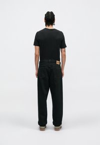 Črna majica s kratkimi rokavi v kombinaciji z temnimi džins hlačami, ki imajo etiketo na pasu in zvite manšete. Model je viden s hrbta.