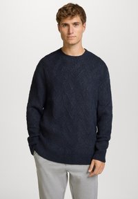 Marineblauwe gebreide trui met een gestructureerd chevronpatroon, ronde hals en geribbelde manchetten. Gecombineerd met lichtgrijze broek voor contrast.