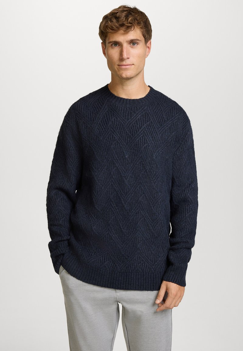 Marineblauwe gebreide trui met een gestructureerd chevronpatroon, ronde hals en geribbelde manchetten. Gecombineerd met lichtgrijze broek voor contrast.