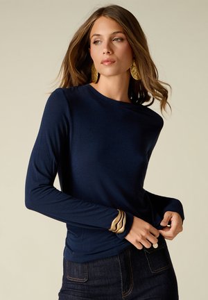 Woman wearing a navy long-sleeve top and dark jeans, accessorized with large gold earrings and a gold bracelet, looking to the side.