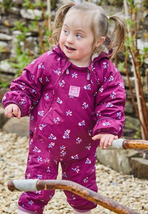 Lila wasserfester Overall mit Blumenmuster, elastischen Bündchen und einem Reißverschluss. Mit einem Logopatch auf der Brust. Kind draußen auf Kies.