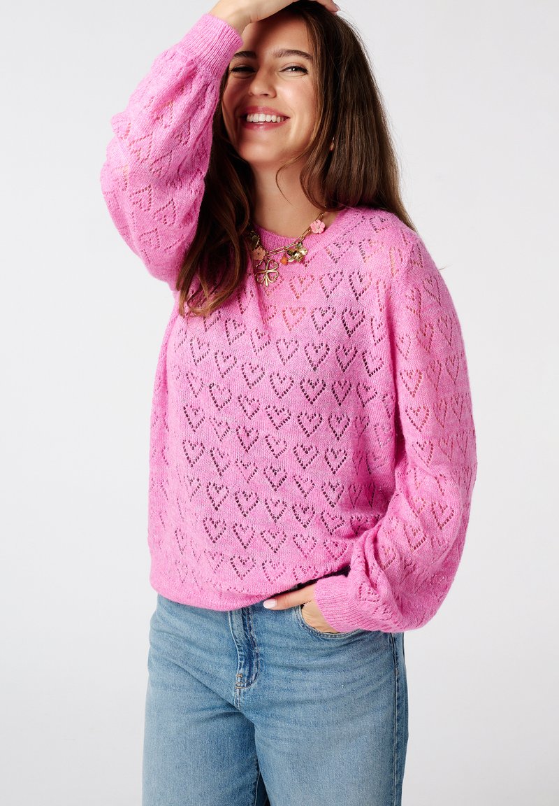 Roze gebreide trui met hartjespatronen, ronde hals en lange mouwen; gecombineerd met een blauwe jeans, wat een casual uitstraling creëert.