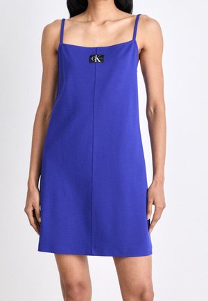 Femme portant une mini-robe sans manches bleu roi avec de fines bretelles et un petit écusson noir Calvin Klein sur la poitrine.
