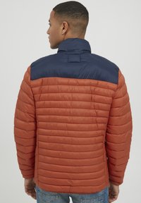 Padded jacket su oranžiniu kūnu ir tamsiai mėlynu apykakle. Qiltižuotas audinys, aukšta apykaklė ir tiesus kirpimas, sukurtas šilumai.