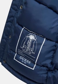 Tamsiai mėlynas pūkinis švarkas su matinio audinio tekstūra, su grafiniu etiketės spaudiniu, vaizduojančiu švarko iliustraciją ir prekės ženklą „GUESS.“