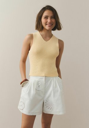 Ung kvinde smilende iført en ærmeløs gul striktop og hvide shorts med blonder i skalmønstre, hænderne i lommerne, foran en ensfarvet baggrund.