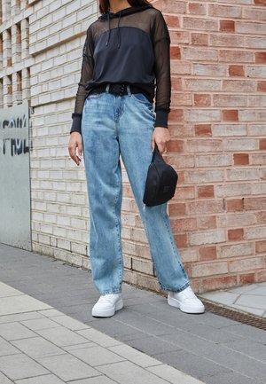 Personne portant un jean bleu clair, des baskets blanches et un haut à capuche noir avec des manches en mesh, tenant un petit sac noir près d’un mur en briques.