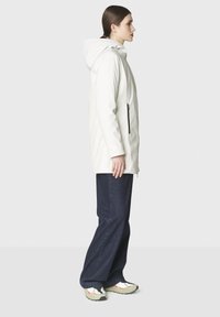 Nainen seisoo sivuttain valkoinen hupullinen takki päällään, tummansiniset leveälahkeiset housut ja valkoiset lenkkarit, joissa on punaisia ja mustia yksityiskohtia.