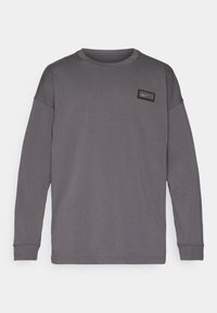 Grå langermet sweatshirt laget av mykt stoff, med rund halsutringning og et rektangulært logo-patch på øvre venstre bryst.