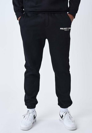 Pantalones deportivos negros de tejido suave, con cintura y puños elásticos, y con el logotipo blanco "PROJECT X PARIS" en la cadera izquierda.