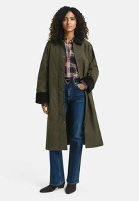 Frau in einem langen olivgrünen Mantel mit schwarzen Ärmeln, einem roten Karoshirt, blauen Jeans und dunkelbraunen Stiefeletten, die mit einer Hand in der Tasche steht.