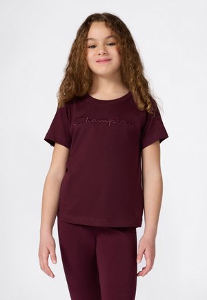 Fată tânără cu păr creț, purtând un tricou și colanți asortați de culoare vișinie închis marca Champion, stând pe un fundal alb simplu.