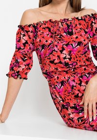 Šaty se spadlými rameny v červeném a černém květinovém vzoru, s krátkými nabíranými rukávy s volánky na okrajích a šňůrkovým výstřihem.