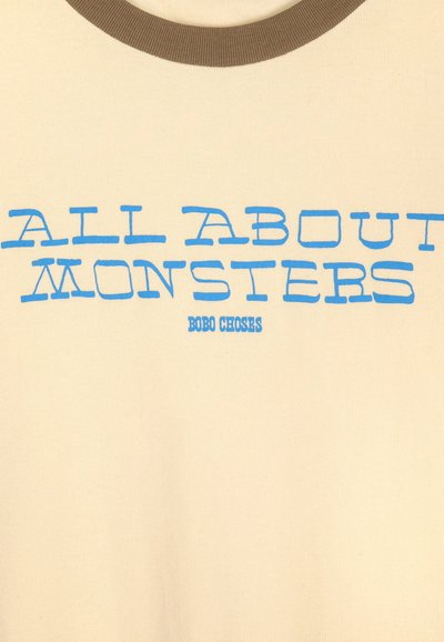 Tissu crème avec col rond marron, texte bleu "ALL ABOUT MONSTERS" et texte bleu plus petit "BOBO CHOSES" en dessous.