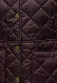 Section de veste matelassée marron foncé avec surpiqûres en losanges, boutons-pression en métal, poches et Barbour brodé sur une poche.