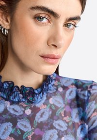 Doorzichtige bloemenblouse met een gerimpelde donkerblauwe halslijn, voorzien van blauwe en paarse bloempatronen op een transparante achtergrond.