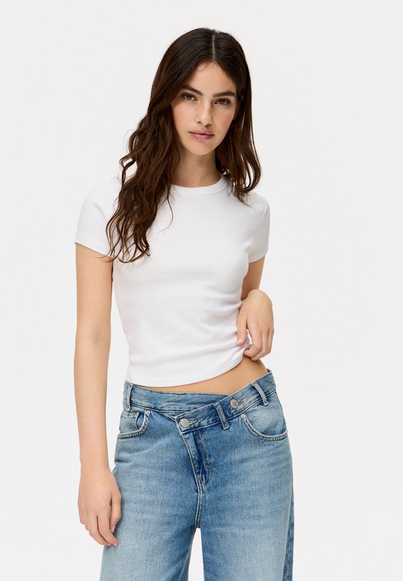 Tricou alb din bumbac, cu mânecă scurtă și decolteu rotund, purtat cu blugi deschis la culoare, cu talie înaltă, care au două buzunare frontale și nasturi.