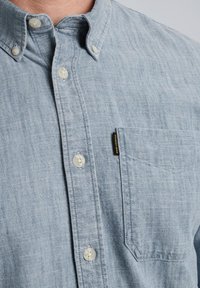 Camisa de botón azul claro hecha de una tela texturizada, con un bolsillo en el pecho y botones de un color claro. Etiqueta pequeña de la marca en el bolsillo.