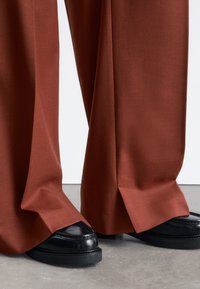 Loose-fitting rust-colored trousers with a smooth texture and side slits, paired with black leather loafers featuring a chunky sole.