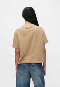 Ανοιχτό καφέ κοντομάνικο πουκάμισο με άνετη εφαρμογή, με στρογγυλή λαιμόκοψη και λεία υφή, σε συνδυασμό με ξεθωριασμένο μπλε τζιν παντελόνι.