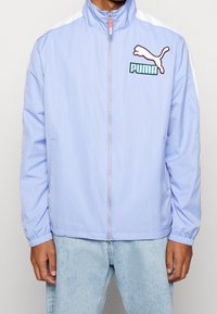 Chaqueta cortavientos azul claro hecha de una tela suave y ligera. Presenta un cuello alto, cierre de cremallera y detalles blancos en las mangas. Logotipo de Puma mostrado.