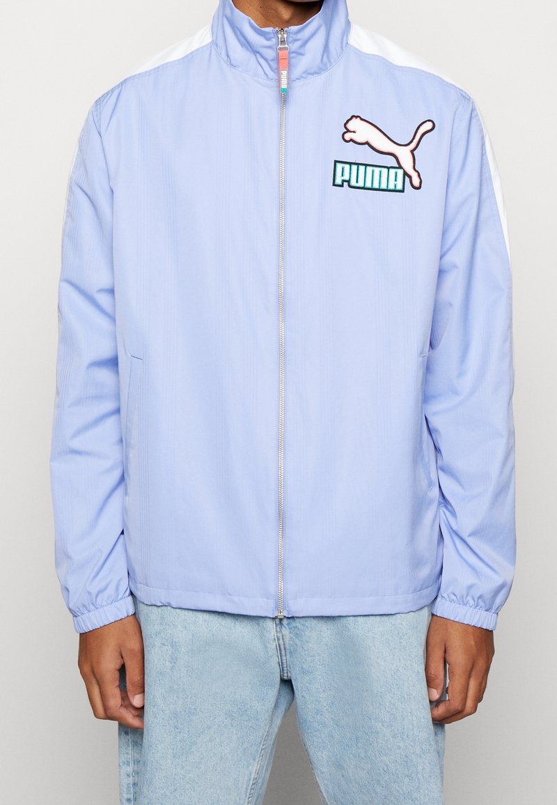 Chaqueta cortavientos azul claro hecha de una tela suave y ligera. Presenta un cuello alto, cierre de cremallera y detalles blancos en las mangas. Logotipo de Puma mostrado.