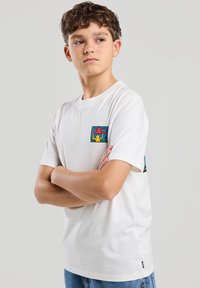 Valkoinen puuvillapaita, jossa on värikkäitä neliölaikkuja rinnassa ja hihoissa, rento istuvuus ja pyöreä kaula-aukko. Rentouttavat siniset farkut.